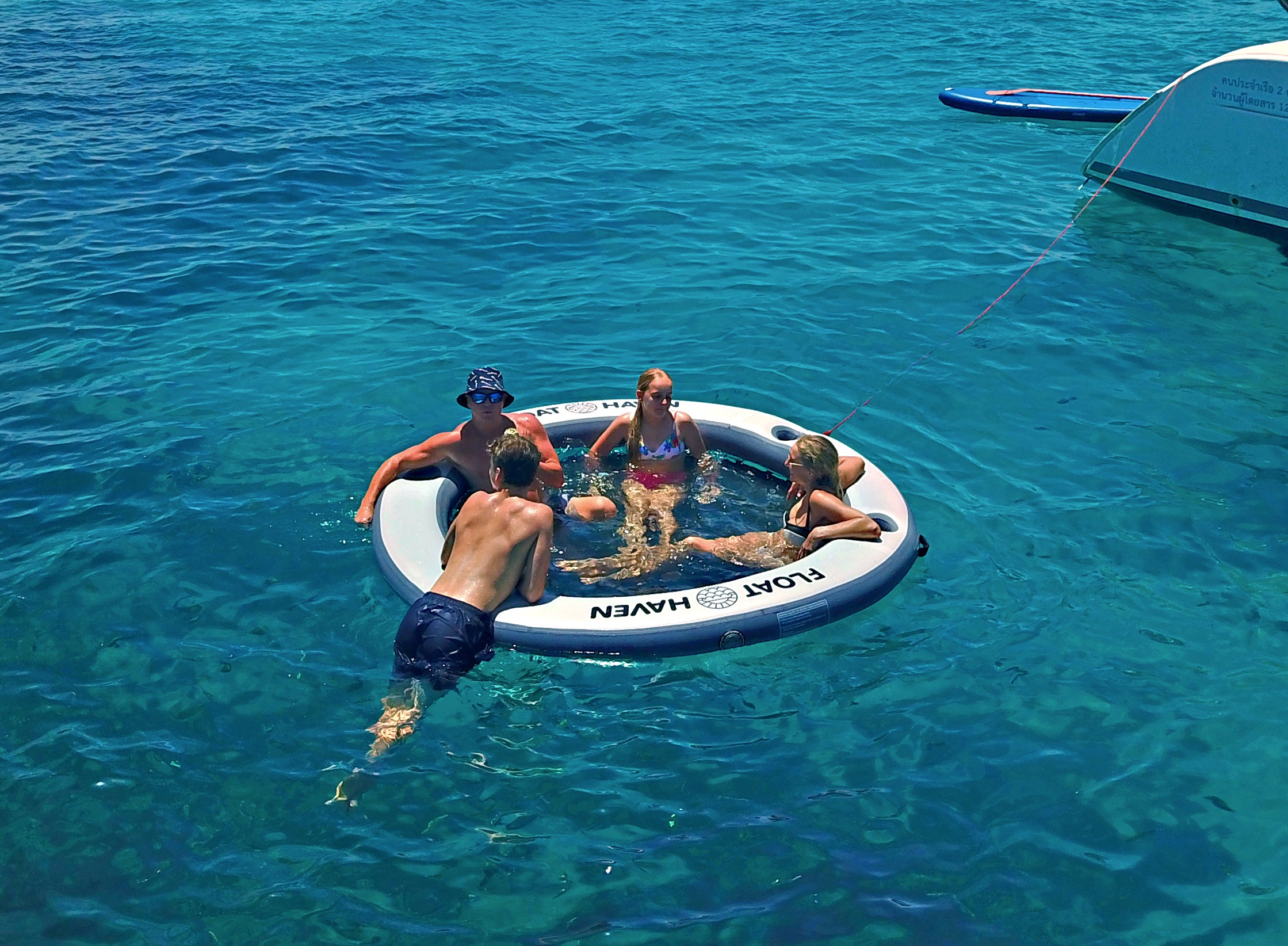Group of friends relaxing on a Float Haven inflatable floating island tied near a boat in clear blue water.