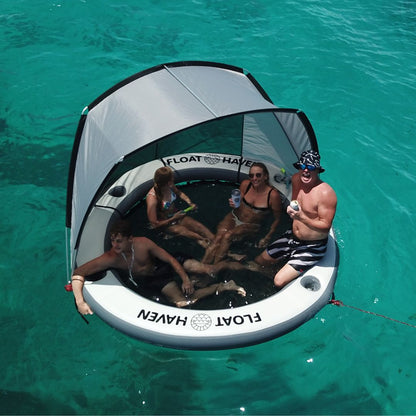 Family relaxing on a Float Haven inflatable floating island with shade canopy and built-in drink holders on turquoise water.